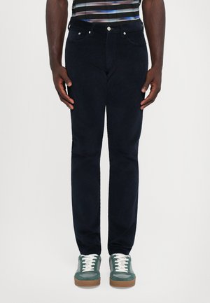 Man med mörkblå kordbyxor, gröna och grå sneakers med vita snörningar, och en randig skjorta, står mot en vit bakgrund.