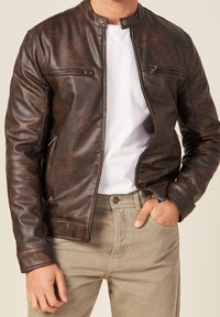 Man die een openbruine lederen jas draagt over een wit T-shirt en beige broek, met zijn rechterhand in de zak en zijn linkerhand ontspannen.