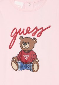 Różowa koszulka z grafiką niedźwiedzia w czerwonym swetrze i niebieskich dżinsach. Słowo "guess" jest wyraźnie widoczne w czerwonym druku powyżej.