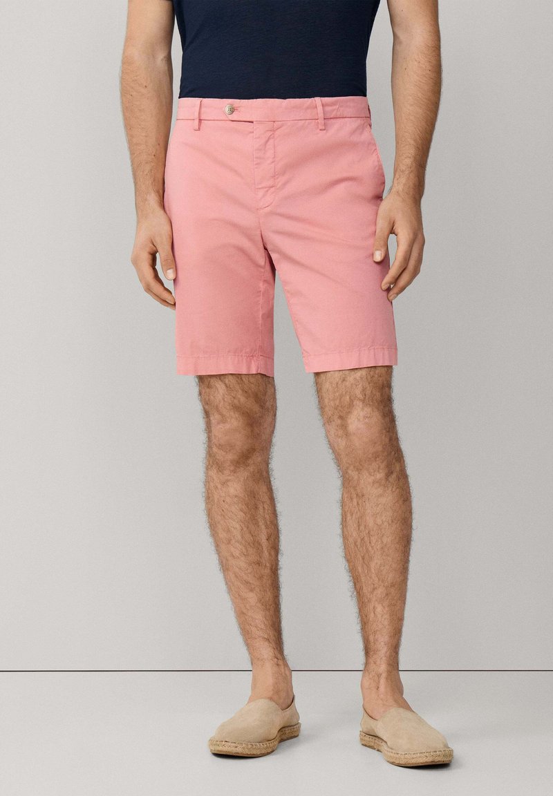 Homme portant un short rose corail, une chemise bleu marine et des chaussures beige sans lacets, debout devant un fond gris clair uni.