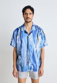 Blue short-sleeve shirt with a wave-like abstract pattern. Features a button-down front, light texture, and contrasting striped shorts.