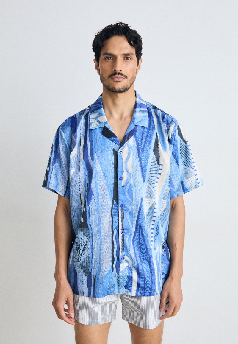 Blue short-sleeve shirt with a wave-like abstract pattern. Features a button-down front, light texture, and contrasting striped shorts.