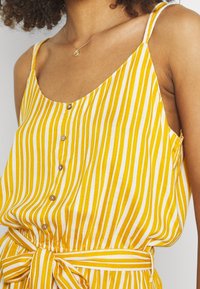 Žena v žluto-bílých pruhovaných bezrukávových šatech se zapínáním na knoflíky vpředu a opaskem svázaným v pase.