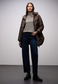Braune Steppjacke mit strukturierter Oberfläche, grauer Strickpullover und dunkelblaue, hoch taillierte Jeans, kombiniert mit schwarzen Stiefeletten.
