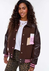 Młoda kobieta z długimi kręconymi włosami, nosząca brązową kurtkę szkolną z różowymi guzikami i spodnie w panterkę, uśmiechająca się do kamery.