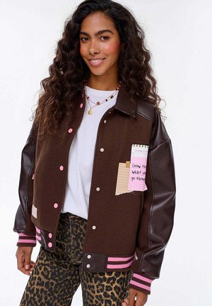 Młoda kobieta z długimi kręconymi włosami, nosząca brązową kurtkę szkolną z różowymi guzikami i spodnie w panterkę, uśmiechająca się do kamery.