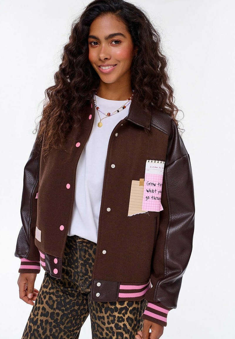 Młoda kobieta z długimi kręconymi włosami, nosząca brązową kurtkę szkolną z różowymi guzikami i spodnie w panterkę, uśmiechająca się do kamery.
