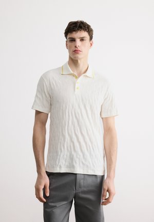 Un jeune homme se tient face à l'avant, portant un polo blanc texturé avec des accents jaunes et un pantalon gris, sur un fond blanc uni.