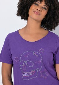 Lilla bomulds T-shirt med korte ærmer, med et flerfarvet broderet kranie- og stjerne-design på fronten. Glat tekstur.