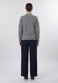 Γυναίκα με τα μαλλιά σε κότσο, φορώντας γκρι ριμπ πλεκτό πουλόβερ, ναυτικό παντελόνι με φαρδιά πόδια και μαύρες γόβες, στέκεται αντικρίζοντας ένα απλό λευκό φόντο.