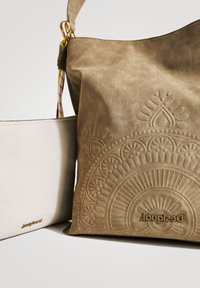 Brązowa torba ze skórzanym wzorem o fakturze z wytłoczonym okrągłym wzorem i logo. Oprócz tego w komplecie kremowa saszetka z złotym detalem logo.