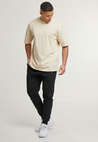 Cremefarbenes, oversized T-Shirt mit kurzen Ärmeln, kombiniert mit schwarzen Jogginghosen. Weiße Sneaker runden das legere Outfit ab.