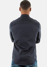 Maglietta a maniche lunghe blu navy con una texture liscia, colletto con bottoni e dettagli sulle spalle, mostrata da dietro su uno sfondo semplice.