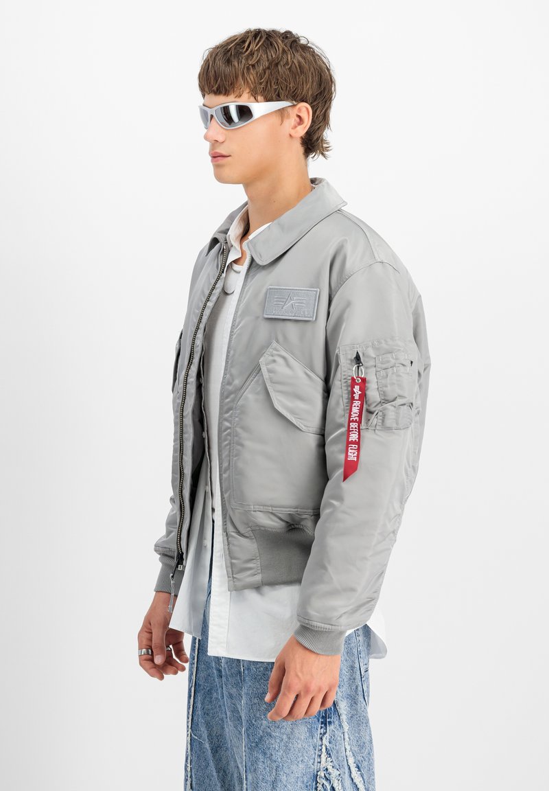Giubbotto bomber grigio con più tasche, polsini a costine e dettaglio con etichetta rossa. Indossato sopra una camicia bianca e pantaloni in denim chiaro, abbinato a occhiali da sole bianchi.