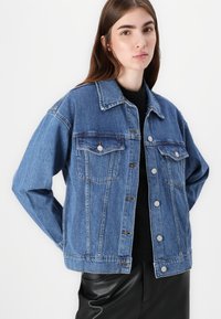 Denim jakna s klasičnim krojem, svetlo modre barve, s prsni žepi in srebrnimi gumbi. Tkanina se zdi teksturirana z rahlim raztegnjenjem.