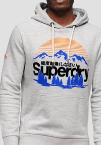 Felpa grigia con cappuccio regolabile, caratterizzata da una grafica colorata di montagne e alberi. Logo "Superdry" in bold con accenti neri e blu.