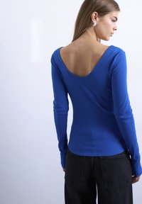 Γυναίκα με μακριά μαλλιά που φοράει ασημένια σκουλαρίκια, μπλε μπλούζα με μακριά μανίκια και βαθύ ανοιχτό ντεκολτέ στην πλάτη, και μαύρο παντελόνι, σε απλό φόντο.