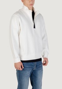 Jersey blanco con cuello de media cremallera, con un acento negro en el cuello y puños acanalados, combinado con unos vaqueros de mezclilla azules.