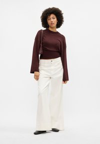 Top longo, cor bordeaux, com cintura ajustada, combinado com calças largas brancas. A modelo segura uma mala de ombro texturada, usando botas pretas.