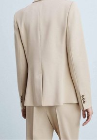 Blazer beige sur mesure avec une coupe structurée, une fente unique au dos, des poignets à deux boutons et une texture de tissu lisse. Pantalon assorti visible.