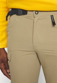 Beiga byxor tillverkade av mjukt tyg, med en cargo-ficka, ett svart justerbart bälte och en knappstängning. Försedda med synlig sömnad.