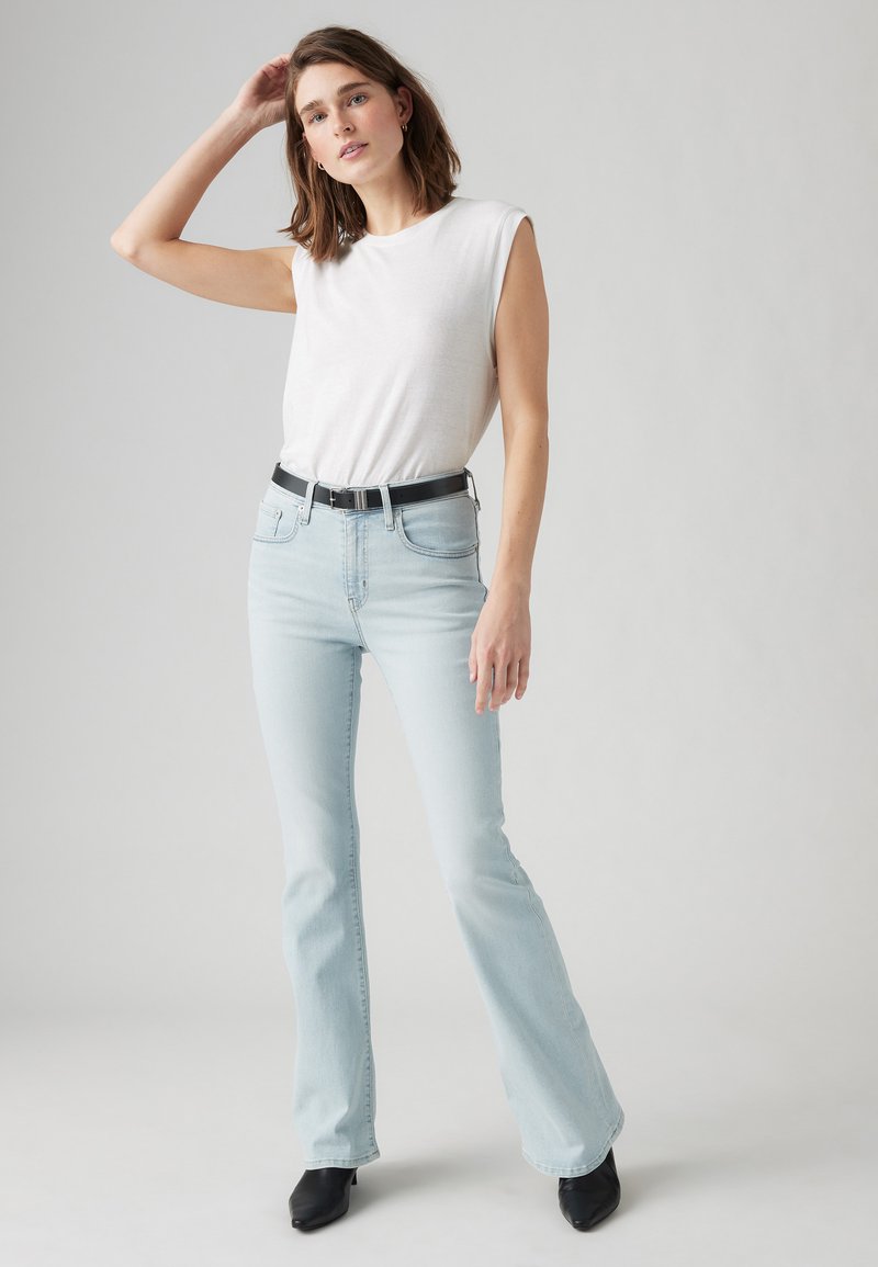 Top blanco sin mangas, jeans acampanados de color azul claro, cinturón negro y botas de tacón negras. Tela suave con textura clásica de denim, diseño minimalista.