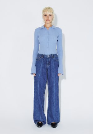 Personne aux cheveux blonds courts portant un pull côtelé boutonné bleu clair, un jean bleu à jambes larges et des chaussures noires pointues, debout contre un mur blanc.