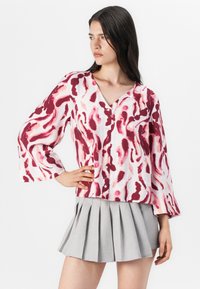 Kvinna med långt hår som bär en mönstrad blus med röda och rosa virvlar, i kombination med en veckad grå kjol, poserar mot en vit bakgrund.