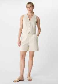 Beige linen vest with five buttons, paired with high-waisted shorts. Textured sandals complete the outfit, emphasizing a cohesive light color palette.