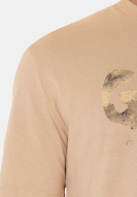 T-shirt in cotone beige con maniche corte e collo rotondo. Grafica testurizzata in oro sul davanti, simile a una lettera o a un logo.