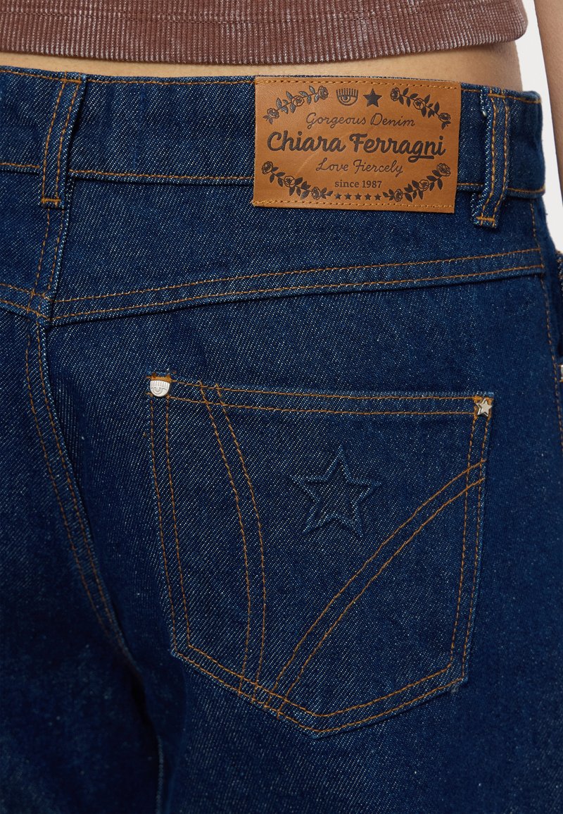 Jean en denim de couleur bleu foncé, avec une étiquette en cuir et une étoile brodée sur la poche arrière. Détails de surpiqûre contrastés.