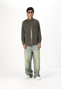 Chemise boutonnée vert olive foncé en tissu doux et texturé, associée à un jean bleu clair délavé avec des jambes larges et des baskets blanches.