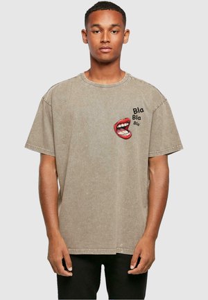 Beige oversized bomull t-skjorte med et rødt og svart grafisk motiv av en munn med teksten "Bla Bla Bla" på venstre brystområde.