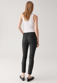 Donna con capelli lunghi che indossa un top bianco senza maniche, jeans neri attillati e ballerine nere, in piedi contro uno sfondo uniforme, girata di spalle.