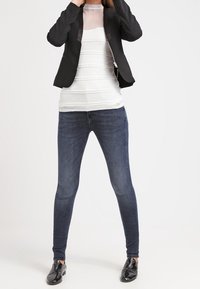 Blazer nero sopra una maglia bianca con texture, abbinato a jeans skinny blu scuro e scarpe slip-on nere. Design semplice e aderente.