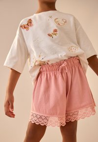 Bílá košile s krátkým rukávem zdobená květinovou a motýlí výšivkou, doplněná o růžové texturované šortky s krajkovým lemem na spodním okraji.