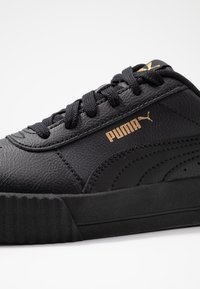 Sneaker en cuir noir avec surface texturée, dotée d'une semelle plate noire, de lacets noirs et d'un logo Puma doré sur le côté.