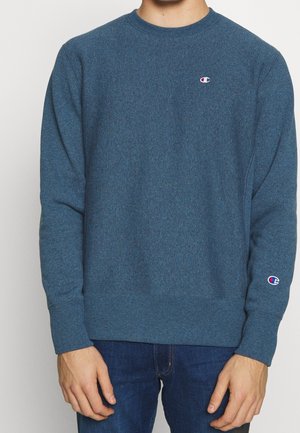 Uomo che indossa una felpa blu girocollo con piccoli loghi Champion sul petto e sulla manica sinistra, abbinata a jeans blu scuro.