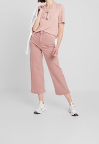 Jasnoróżowy krótki top z krótkim rękawem i wiązaniem przy szyi, zestawiony z luźnymi jasnoróżowymi szerokimi spodniami i białymi trampkami.