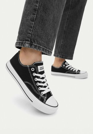 Osoba nosząca czarne niskie trampki z płótna z białymi sznurowadłami i podeszwami, w połączeniu z ciemnoszarymi przetartymi jeansami o długości 7/8 na białym tle.