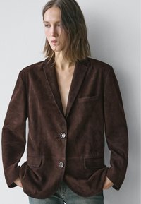 Brązowa zamszowa marynarka z klapą z wycięciem, dwoma przednimi kieszeniami, jednorzędowym zapięciem i kontrastującymi jasnymi guzikami.