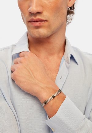 Pulsera de acero con diseño de alambre retorcido y acentos negros. Lleva puesta en una muñeca, combinada con una camisa texturizada de color azul claro.