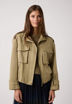 Beige Kurzjacke mit zwei vorderen Taschen, Knopfmanschetten und einem Reißverschluss. Getragen über einem grünen Strickpullover mit einem plissierten dunkelbraunen Rock.
