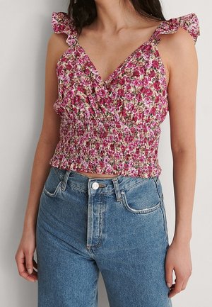 Kobieta ubrana w bezrękawnikowy kwiatowy crop top z falbankowymi ramiączkami i niebieskie jeansy z wysokim stanem, stoi przy gładkiej ścianie.