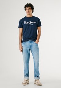 Marineblå t-shirt med hvid "Pepe Jeans London" logo, parret med lyseblå jeans og beige sneakers. Enkel design, bomuldsstof, afslappet pasform.