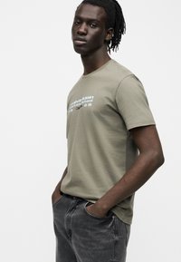 Άνδρας με dreadlocks που φοράει ένα χακί t-shirt Calvin Klein και σκούρα γκρι τζιν, στέκεται με τα χέρια στις τσέπες μπροστά σε λευκό φόντο.