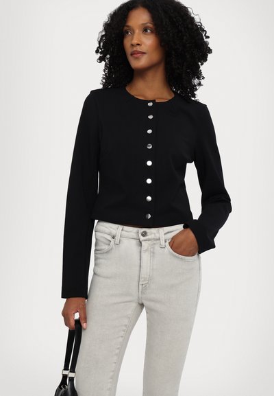 Czarna krótka kurtka z gładkiego materiału, z okrągłym dekoltem i sześcioma dużymi srebrnymi guzikami z przodu. Połączona z jasnymi dżinsami.