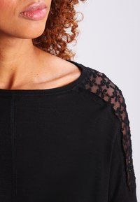 Zwarte top met een losse pasvorm, geribde textuur en doorschijnende kanten schouders versierd met kleine bloemmotieven. Ontwerp met een ronde hals.