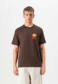 Ung mand iført en brun T-shirt med et farverigt burgergrafik og teksten "Sun Out Buns Out", kombineret med lysebeige bukser.