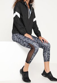 Czarna lekka bluza z białymi paskami, zestawiona z czarnymi legginsami z wzorem i siateczkowymi elementami. Czarne sportowe buty.